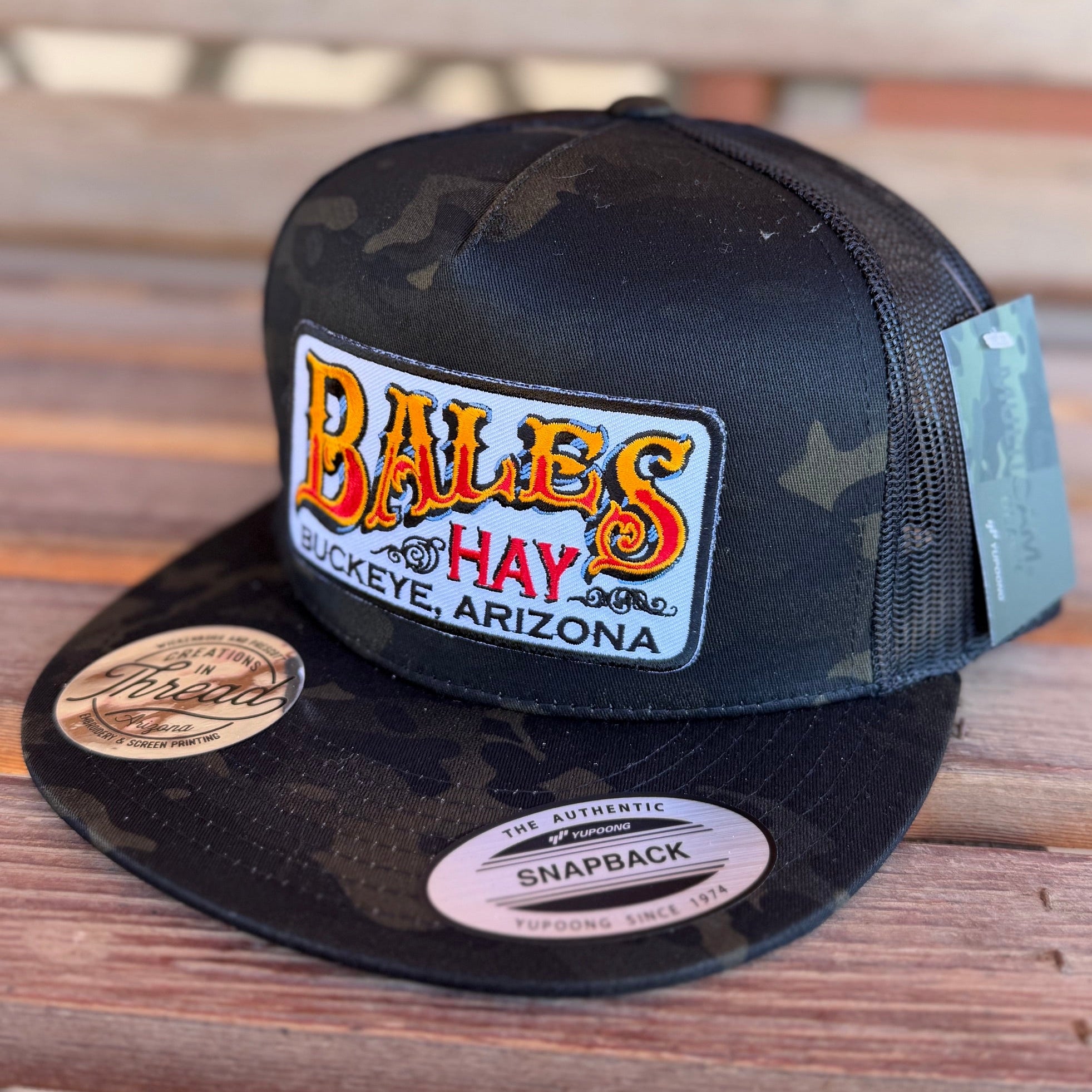 Black and camouflage snapback cap with 'Bales Hay' logo on a wooden surface
