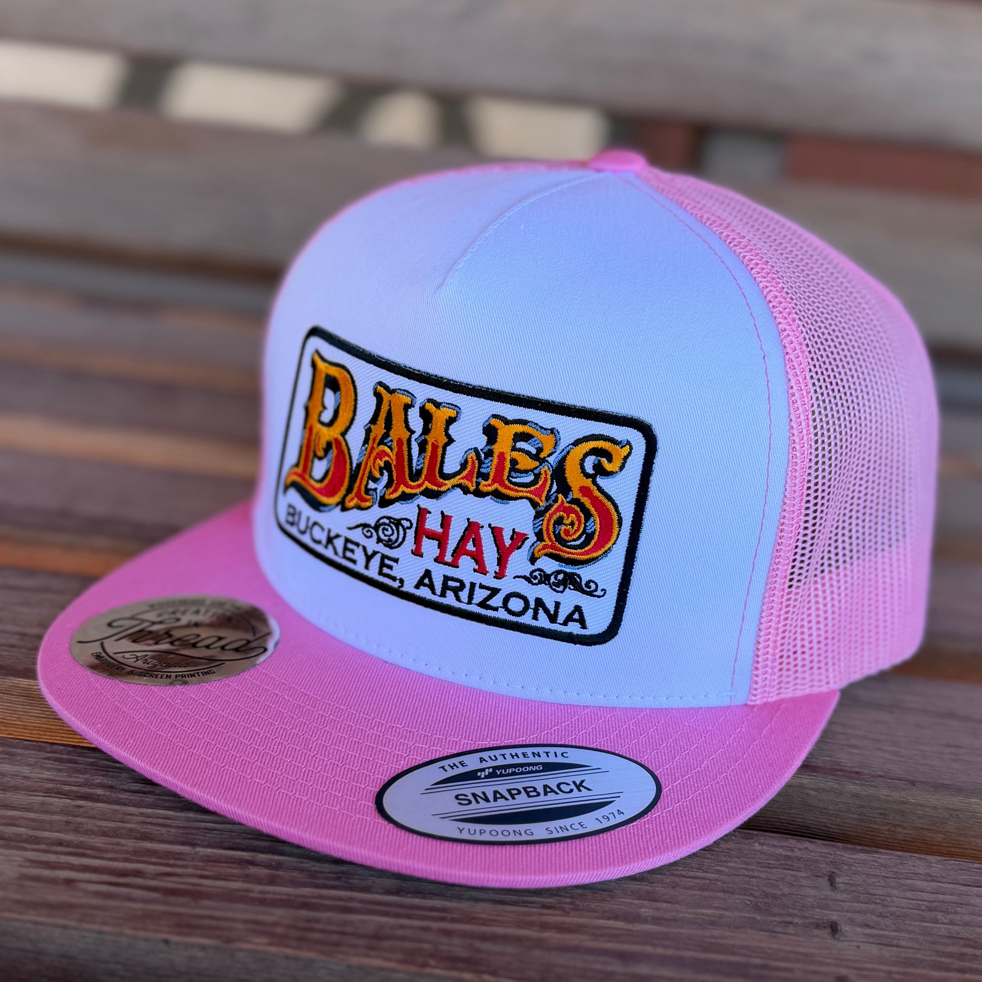 Pink and white cap with 'Bales Hay' logo on a wooden surface